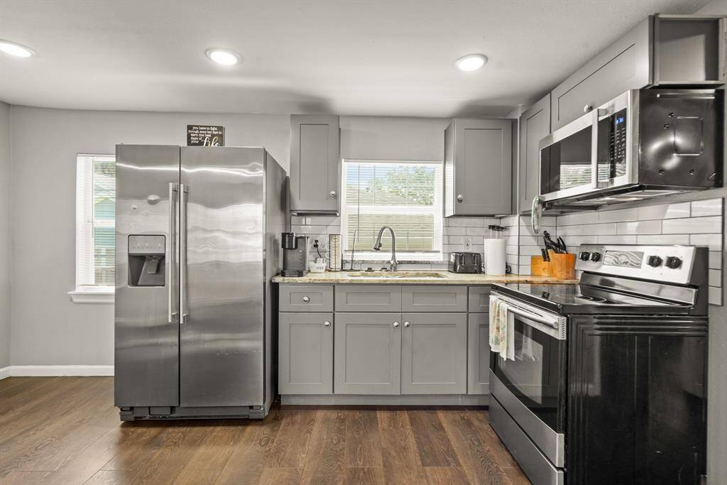 This bedroom features a modern design with neutral tones, hardwood flooring, and ample natural light from two windows. It includes a ceiling fan, a wall-mounted TV, a comfortable bed, and a small desk area, ideal for relaxation and work.