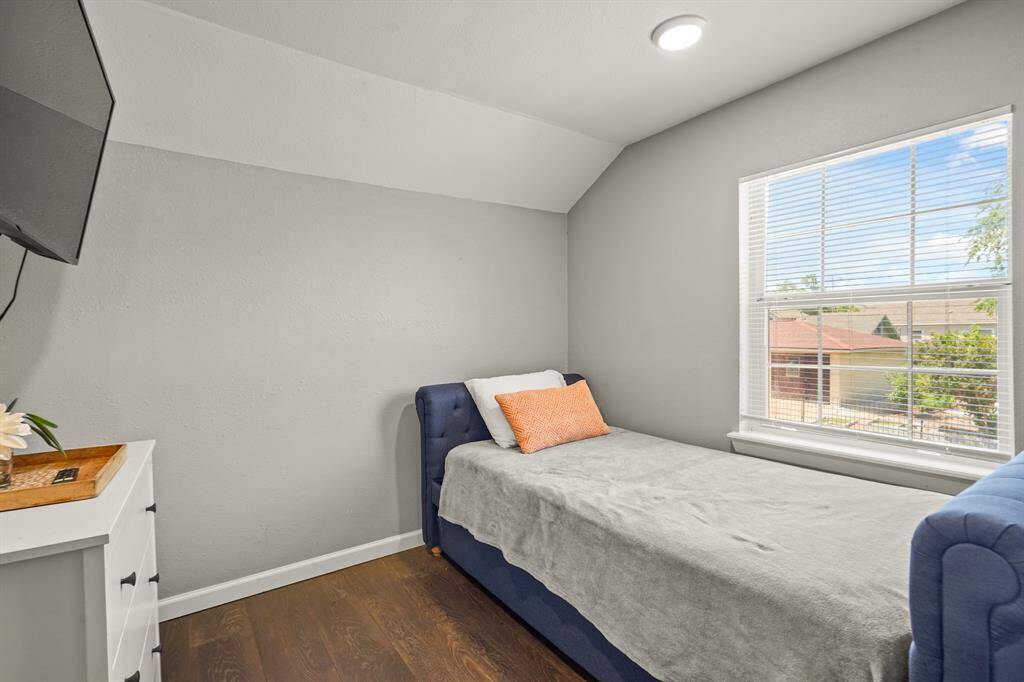 This bedroom features a cozy layout with a ceiling fan, neutral walls, and hardwood floors. It includes a wall-mounted TV, a small dresser with a mirror, and a window for natural light. The room connects to another space through an open door, enhancing its accessibility.
