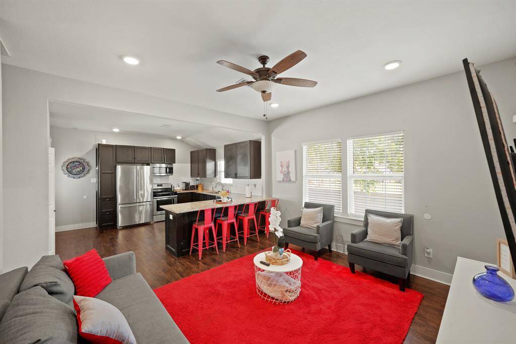 Bright and modern living room with neutral walls, featuring a ceiling fan, recessed lighting, and ample natural light from large windows. It includes a comfortable gray sofa with red accents, two armchairs, a wall-mounted TV, and a stylish clock. The red area rug adds a pop of color, complementing the cozy and inviting atmosphere.