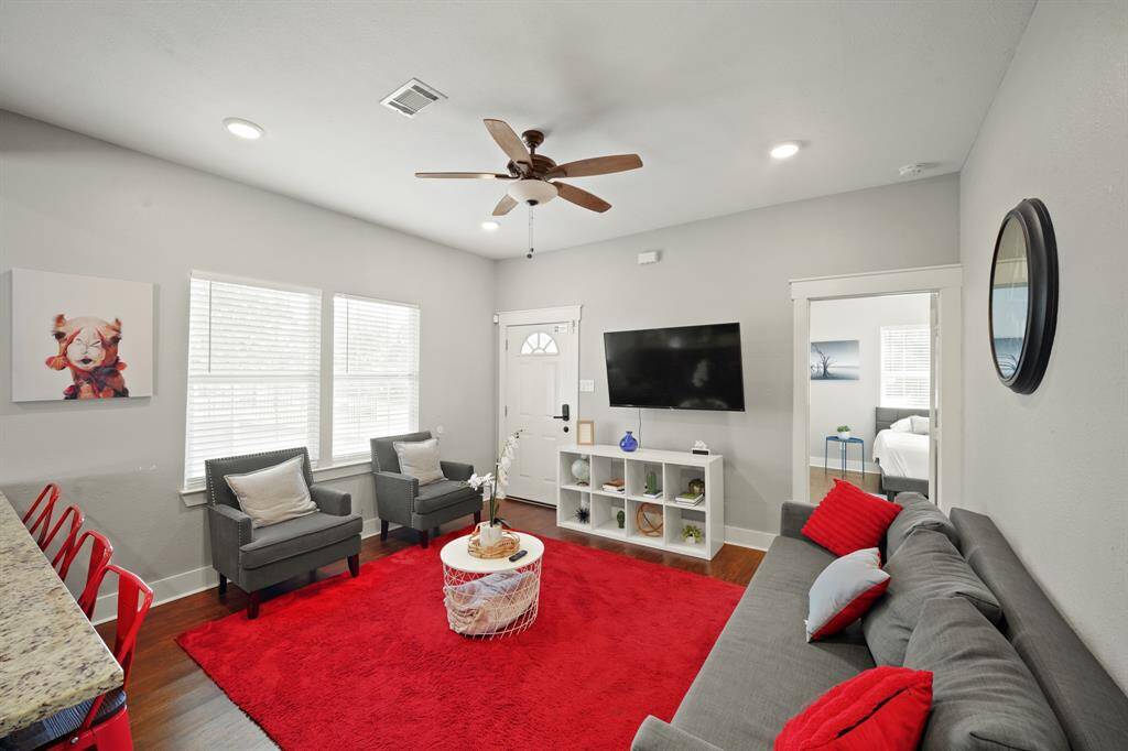This modern living area features an open-concept design with a cozy gray sofa, vibrant red accents, and a ceiling fan. The adjoining kitchen boasts dark cabinetry, stainless steel appliances, and a spacious breakfast bar with red stools, offering a stylish and functional space.