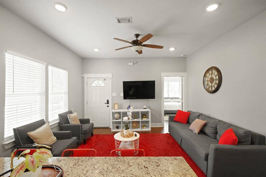 This bright and open living area features hardwood looking floors, a modern kitchen with dark cabinetry and stainless steel appliances, and a cozy seating space. A red rug and bar stools add vibrant accents, while large windows provide ample natural light.