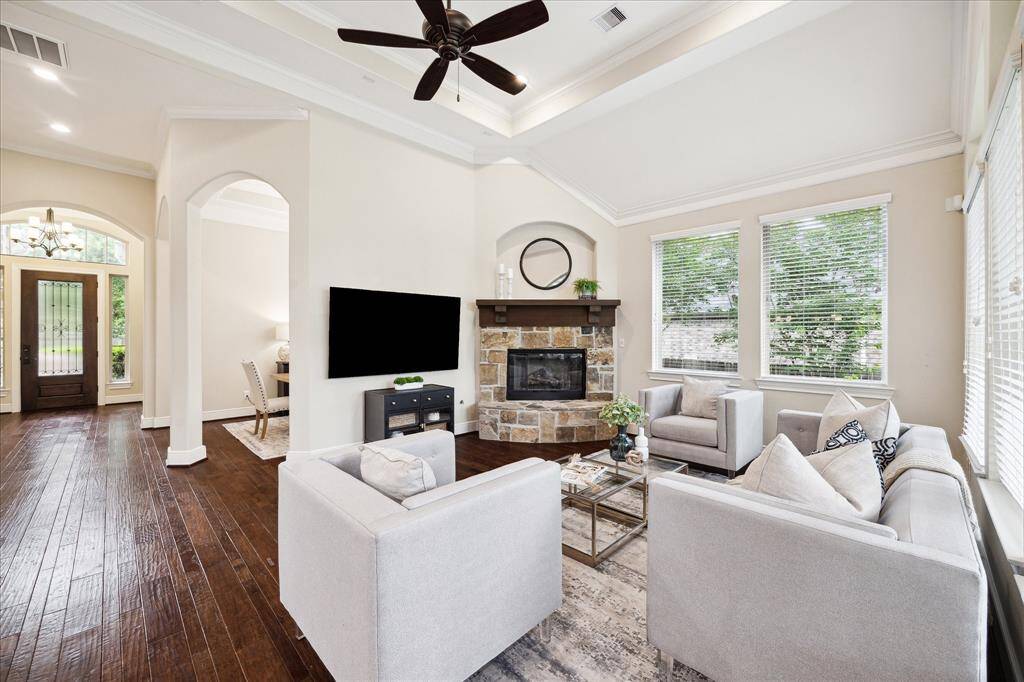 This inviting living room features high ceilings, large windows for natural light, and a cozy stone fireplace. The open layout connects to the kitchen, making for a wonderful family environment or for entertaining.  The dark hardwood floors add a nice touch of elegance.