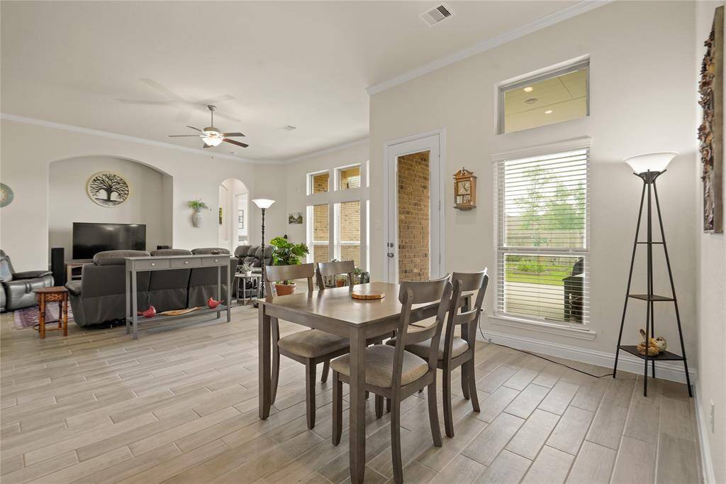 This photo shows a bright, open-concept living and breakfast area with light wood-look tile flooring. The space features a comfortable seating area with a TV, a dining table, large windows for natural light, and a door leading to a patio.