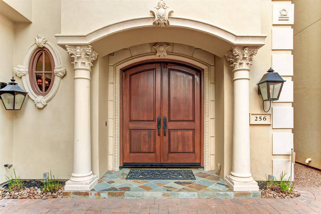 This elegant front entrance features a grand wooden double door framed by decorative columns and an arched stonework, complemented by gas lanterns and a small oval window. The entryway is accented with a stone-tiled step, adding a touch of sophistication to the home's facade.