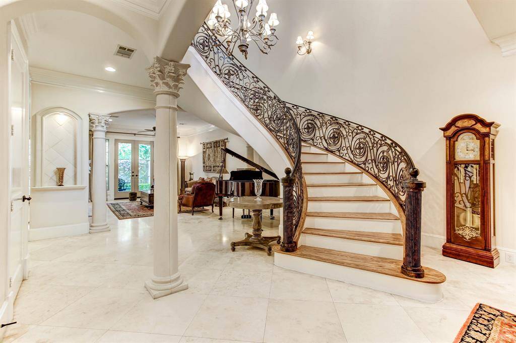 This elegant entryway features a grand staircase with ornate iron railings, a classic chandelier, and travertine flooring. The space is open and airy, leading to a living area with space for a grand piano.