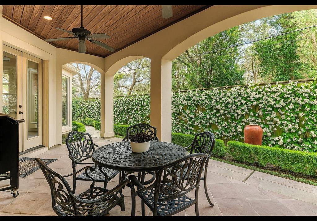 This photo showcases a charming covered patio with a ceiling fan, elegant stone flooring, and arched openings leading to a private garden area with lush greenery and a vine-covered fence, perfect for outdoor relaxation and dining. Note, the jasmine on the fence was done virtually.  The fence was recently replaced with a new one, this is what you could do.