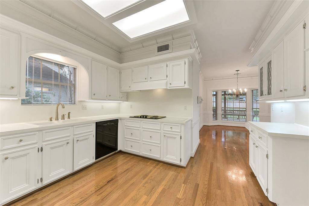 This photo showcases a spacious, bright kitchen with white cabinetry and wood floors. It features modern lighting, a large window, and an open layout leading to a dining area with a view of the backyard.