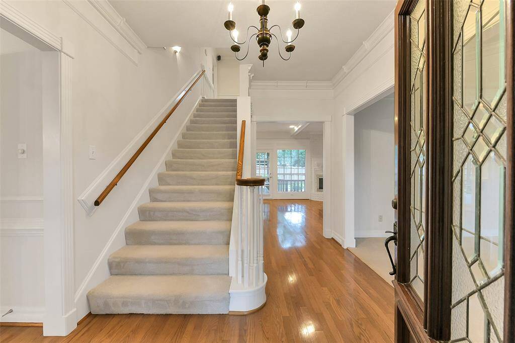 This inviting entryway features a carpeted staircase with elegant wooden handrails, leading to an upper floor. The space is well-lit with a classic chandelier, and the hardwood flooring extends into a bright room with large windows, creating a warm and welcoming atmosphere.