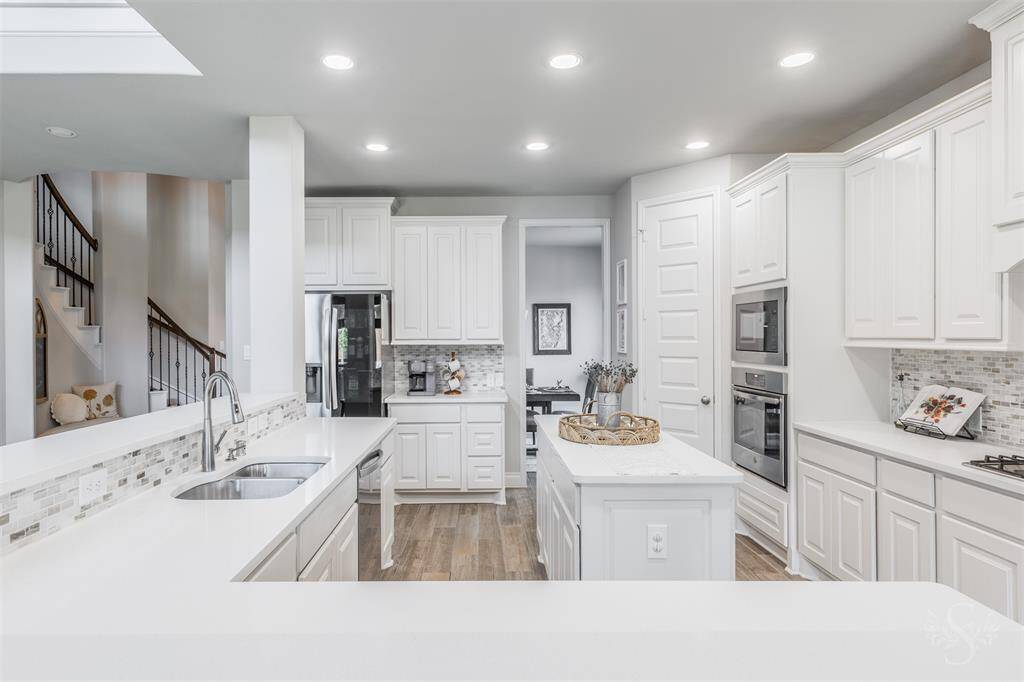 The gourmet kitchen features crisp white cabinetry, stainless steel appliances, and a striking tile backsplash. A long breakfast bar offers ample seating and opens to the living area, making it the heart of the home.