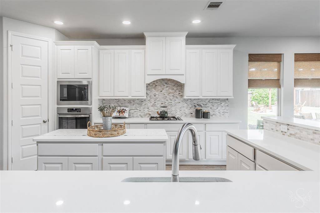 The gourmet kitchen features a gas cooktop, center island, and sleek white cabinetry with detailed molding. A mosaic tile backsplash and expansive counter space make this a chef’s dream.