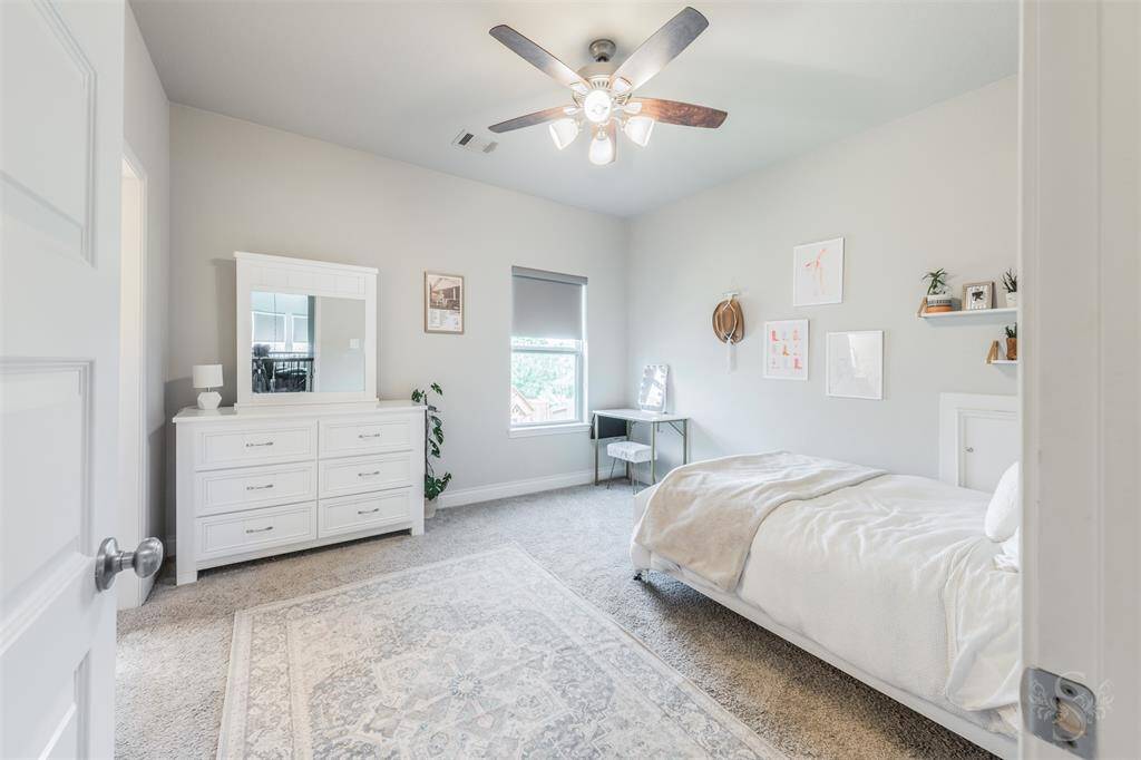 Spacious secondary bedroom features soft carpeting, neutral tones, and a ceiling fan, with space for a bed, dresser, and a cozy desk nook by the window. Natural light fills the room, creating a bright and inviting retreat.