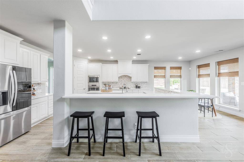 The gourmet kitchen features crisp white cabinetry, stainless steel appliances, and a striking tile backsplash. A long breakfast bar offers ample seating and opens to the living area, making it the heart of the home.
