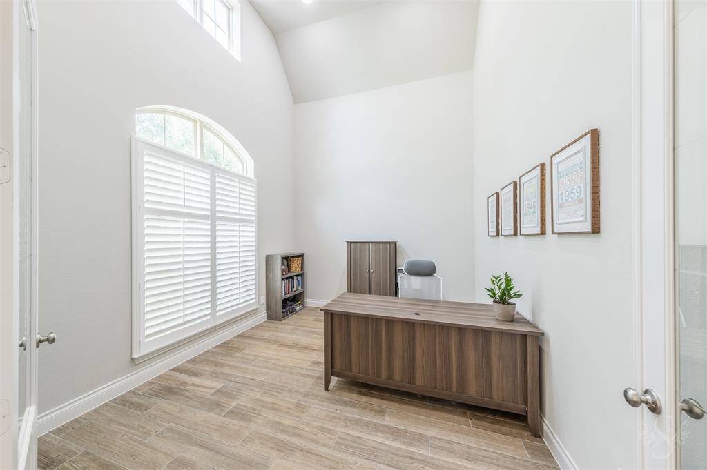 The private study features double glass doors, soaring ceilings, and wood-look tile flooring for a refined yet comfortable workspace. Tall windows with plantation shutters fill the room with natural light, making it ideal for work or quiet reflection.