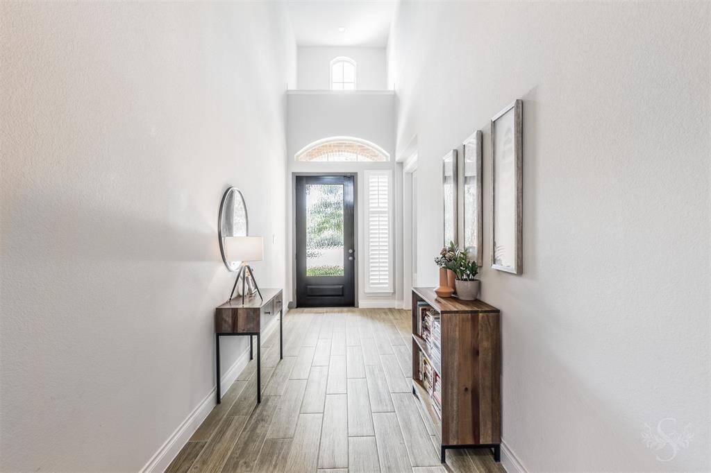 This inviting entryway features tall ceilings, transom windows, and a beautiful glass-panel front door that floods the space with natural light. The elegant wood-look tile flooring continues throughout, setting a warm, upscale tone.