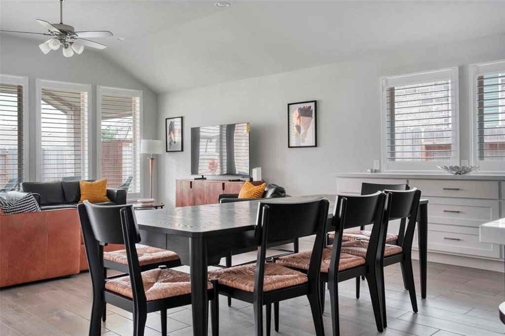 This bright, open-plan living and dining area features large windows with shutters, modern furnishings, and a ceiling fan. The space includes stylish seating, a dining table, and a TV, creating a comfortable and inviting environment.