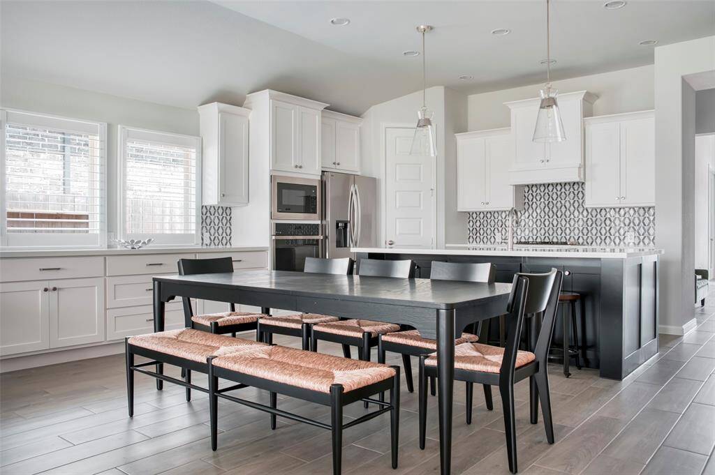 This modern kitchen features sleek white cabinetry, a stylish patterned backsplash, and stainless steel appliances. It includes a spacious island with pendant lighting, and a dining area with a large table and seating for six. The room is bright and airy with ample natural light from the windows.