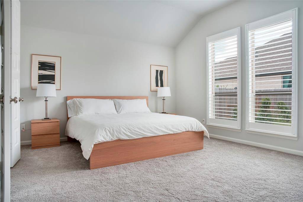 This bedroom features a spacious layout with a large bed, neutral carpeting, and ample natural light from tall windows with blinds. Simple decor and matching wooden nightstands create a modern, cozy atmosphere.