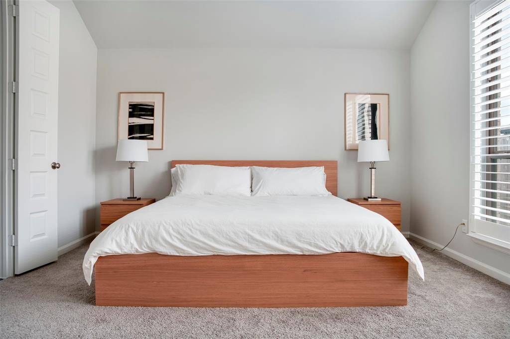This bedroom features a modern, minimalist design with a comfortable bed, wooden side tables, and matching lamps. The room is bright with neutral walls and carpet, enhanced by natural light from large windows with blinds.