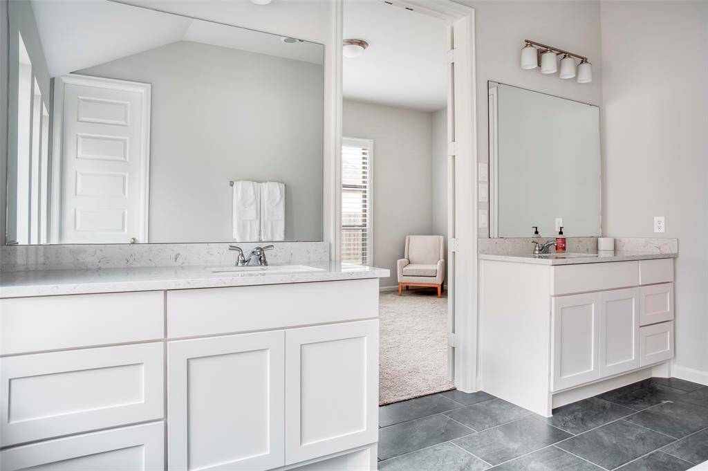 This photo showcases a modern bathroom with dual sinks, white cabinetry, and a large mirror. It features sleek gray tile flooring and an adjoining carpeted room with natural light, creating a bright and inviting space.