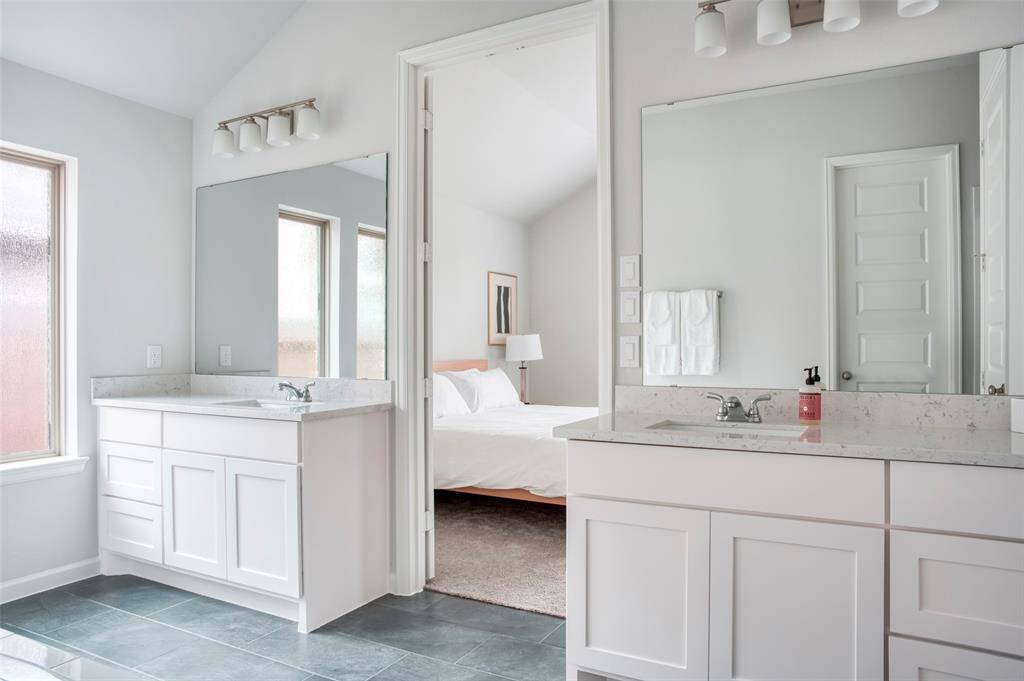 The photo shows a bright, modern bathroom with dual sinks, large mirrors, and sleek white cabinetry. It connects to a bedroom with a glimpse of a bed and soft carpeting, creating a seamless, elegant space.