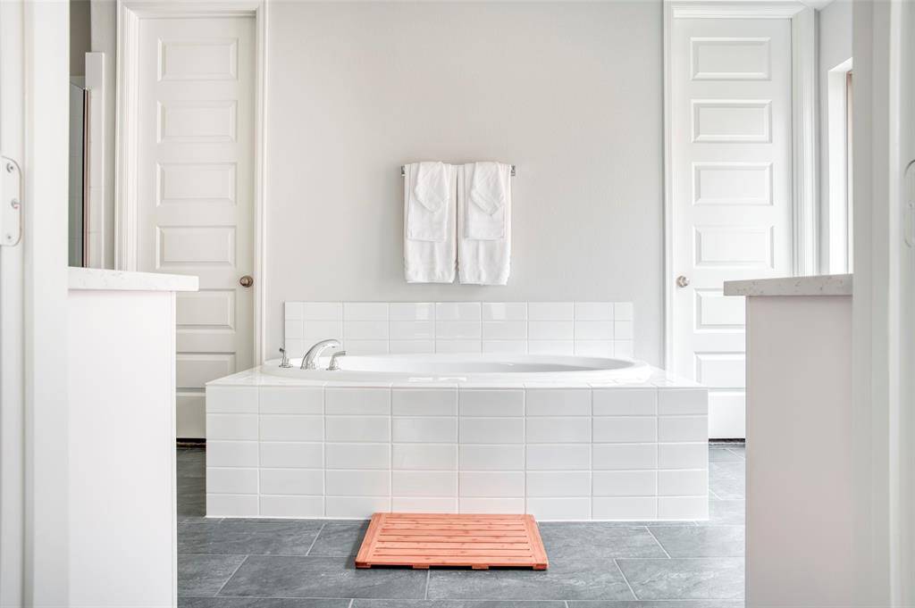 This bathroom features a sleek, modern design with a large white tiled bathtub at the center, flanked by two closed doors, gray tile flooring, and a minimalist palette.