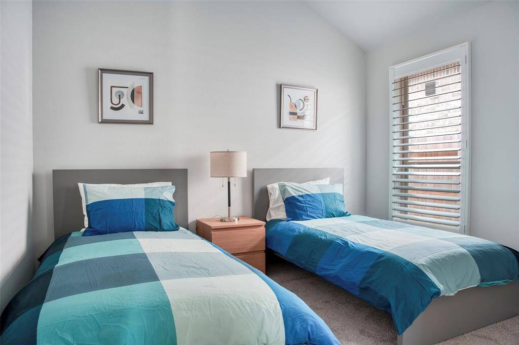 This cozy bedroom features two twin beds with modern blue bedding, a shared nightstand with a lamp, and a window with blinds, allowing natural light to brighten the space. Minimalist artwork decorates the walls, adding a contemporary touch.