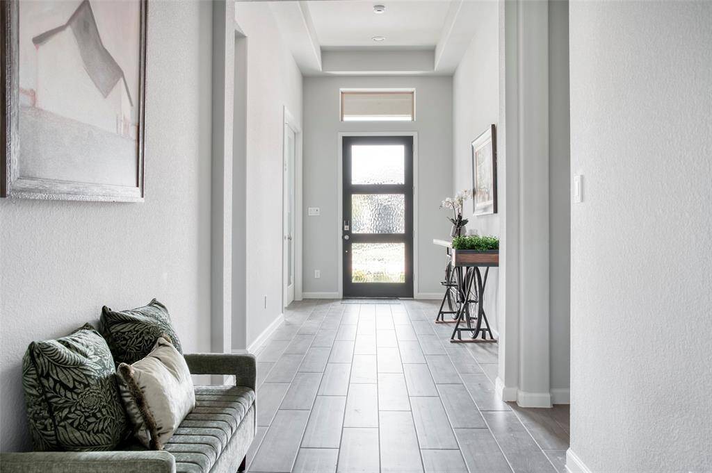 The photo showcases a bright, welcoming hallway with modern tile flooring, neutral walls, and a glass-paneled front door. It features tasteful decor, including a bench with cushions and a console table with plants and artwork, creating an inviting entryway.