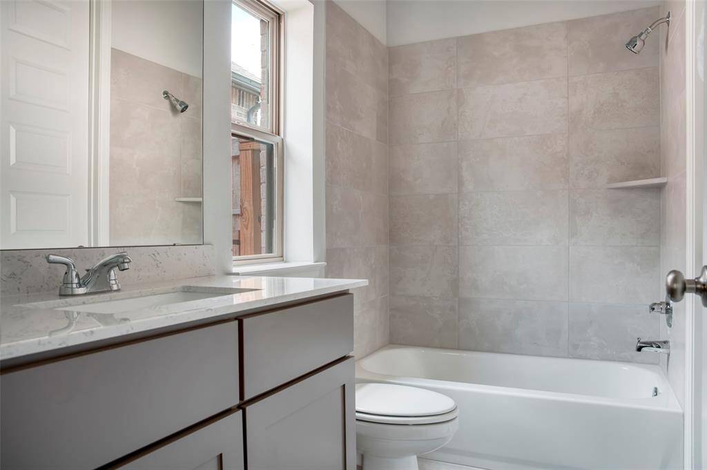 This photo features a modern bathroom with a sleek, tiled bathtub and shower combination. It has a large window allowing natural light, a stylish vanity with a marble countertop, and contemporary fixtures. The neutral tones create a clean, spacious feel.