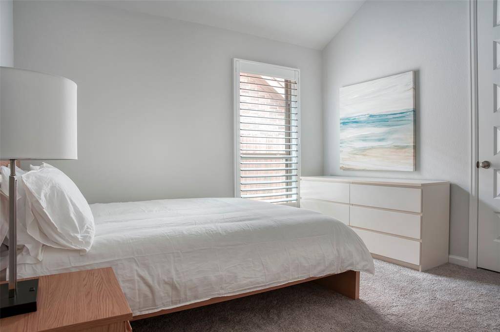 This bedroom is bright and airy, featuring a large window with blinds, a cozy bed with white linens, a modern dresser, and a calming ocean-themed artwork. The neutral color palette creates a serene atmosphere.
