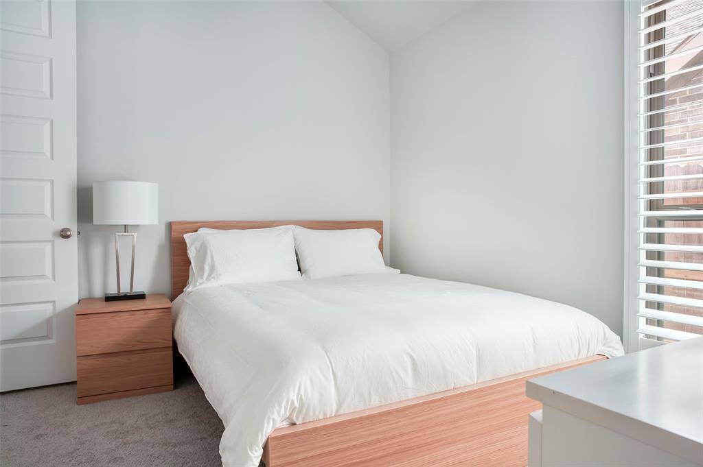This bedroom features a modern, minimalist design with a comfortable bed, neutral tones, and ample natural light from large windows. The space includes a wooden nightstand with a sleek lamp, creating a cozy and inviting atmosphere.
