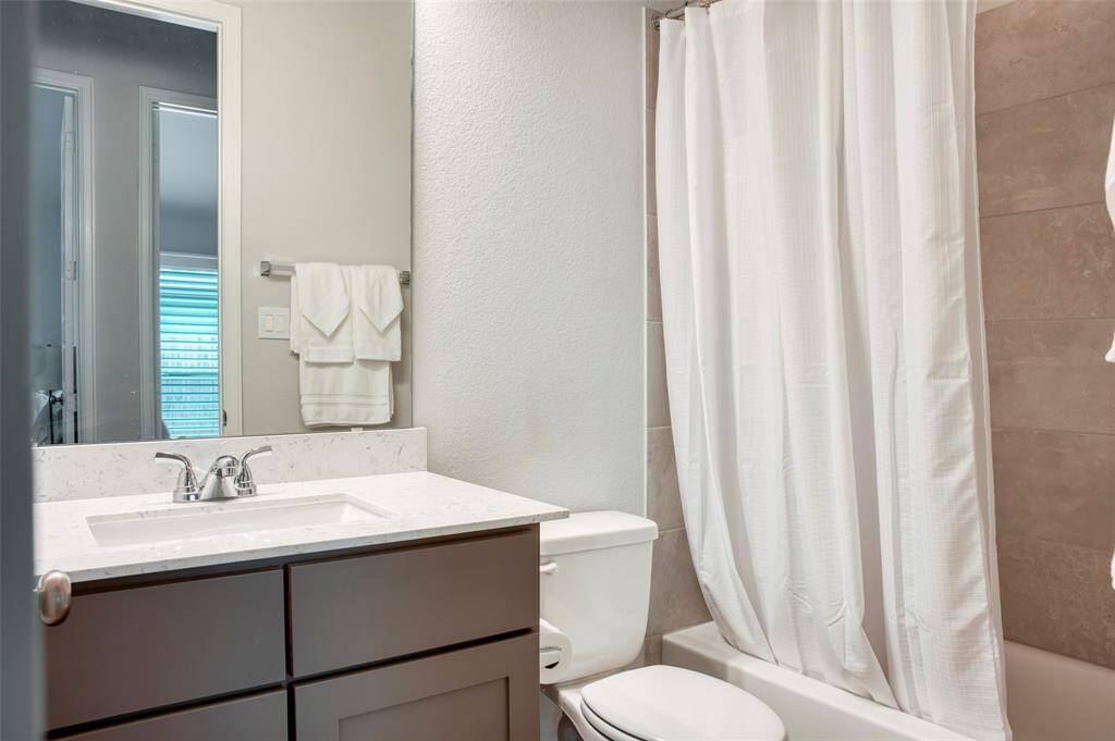 This bathroom features a modern vanity with a marble countertop, a sleek faucet, and ample cabinet space. It includes a toilet and a bathtub with a white shower curtain. The light-colored tiles and neutral tones create a clean, bright atmosphere.