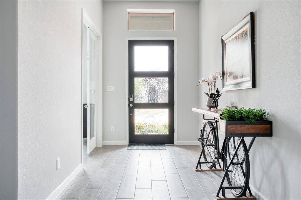 This photo showcases a bright and welcoming entryway with modern design elements. It features a glass-paneled front door, light-colored walls, and wood-look tile flooring. A unique console table doubles as a planter, adding a touch of greenery, and a large framed artwork adorns the wall. The space is both stylish and functional, offering a warm first impression.