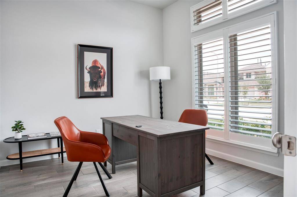 This bright and inviting home office features large windows with shutters, a sturdy wooden desk, and stylish orange chairs. A floor lamp and wall art add charm, while the view outside offers a glimpse of a well-kept neighborhood.