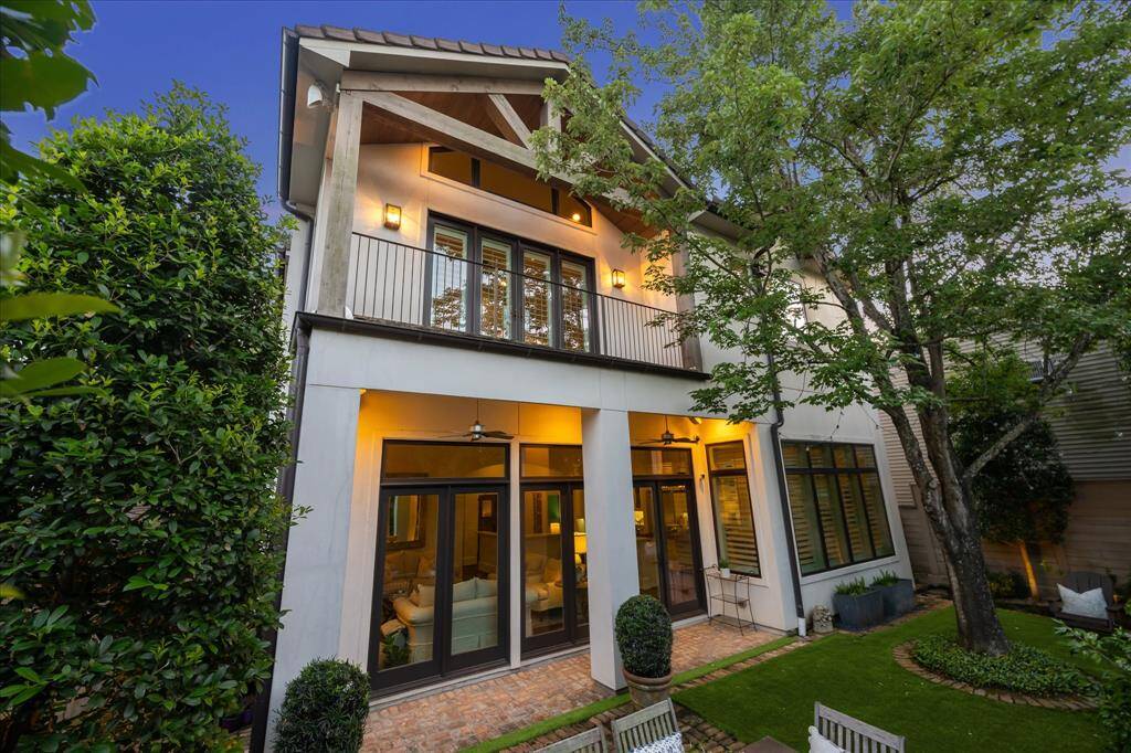 Striking rear elevation with expansive windows, upper balcony, and covered brick terrace shaded by mature trees.