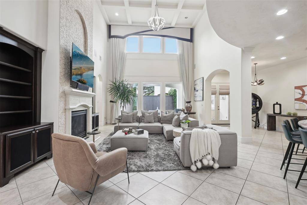 This spacious living room features high ceilings, a large chandelier, and a modern open layout. It includes a cozy fireplace, and ample natural light from tall windows.