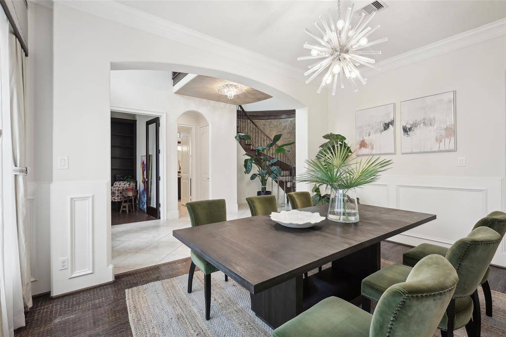 This elegant dining room features a modern chandelier, green velvet chairs, and a dark wood table. It opens to a hallway with arches and a staircase, enhancing the space's openness and sophistication.