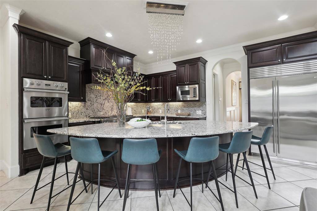 This modern kitchen features dark wood cabinetry, a large granite island with seating, stainless steel appliances, and a stylish chandelier. It's spacious and ideal for entertaining.