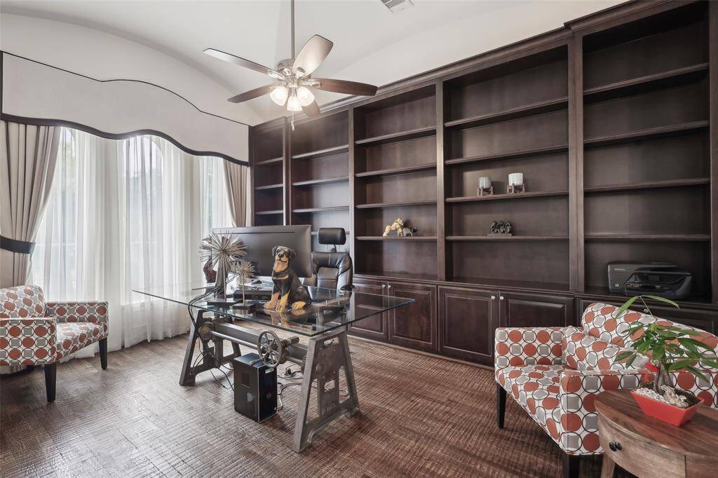 This home office features a high ceiling and large window with elegant curtains, filling the room with natural light. It includes built-in dark wood shelving, a modern glass desk, and comfortable patterned chairs, providing a stylish and functional workspace.