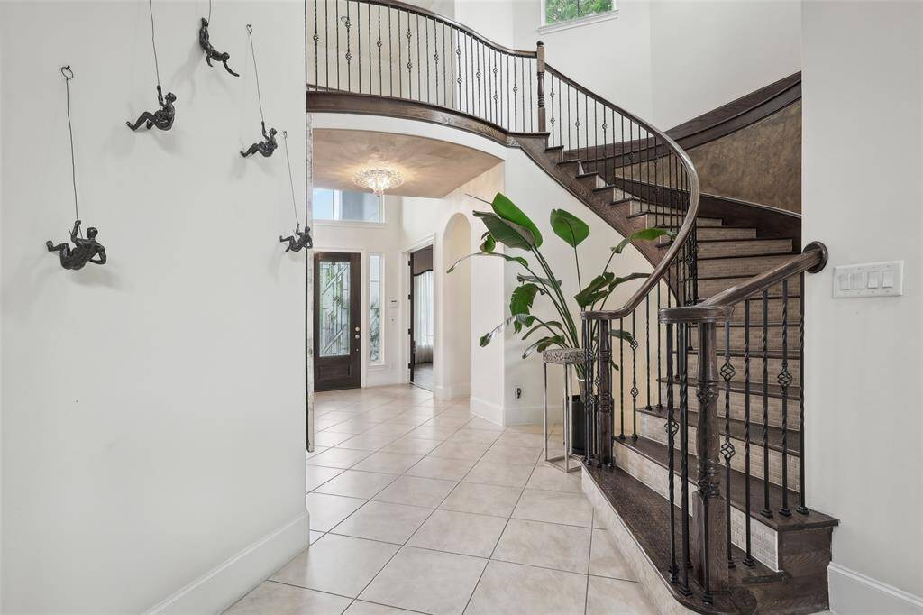 This elegant entryway features a grand, curved staircase with wrought iron railings, large tile flooring, and a modern chandelier. Decorative wall art and a stylish plant add character, while high ceilings enhance the spacious feel.