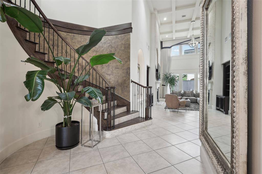 This elegant home features a grand entryway with a stunning curved staircase and modern iron railing. The space is bright and open, with high ceilings and large windows in the living area. A large mirror and lush plant add sophistication, while the neutral tile flooring complements the modern design.