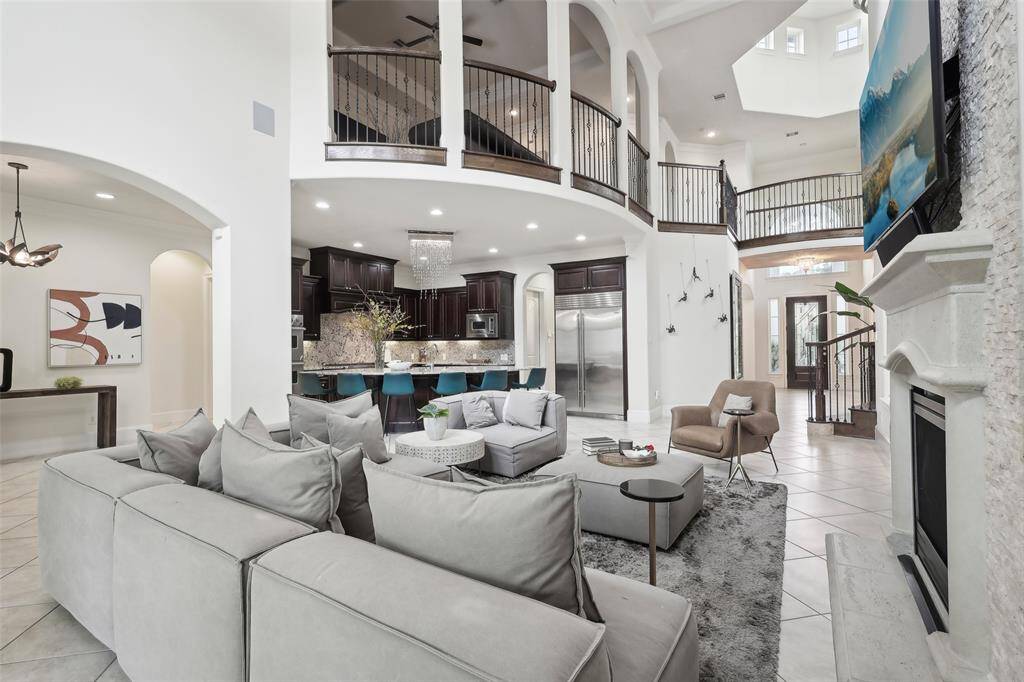 This spacious living area features high ceilings, elegant arches, and a modern open floor plan. The room is anchored by a cozy fireplace, with a view of the stylish kitchen featuring dark cabinetry, stainless steel appliances, and a breakfast bar. The second-floor balcony overlooks the space, adding a touch of grandeur.