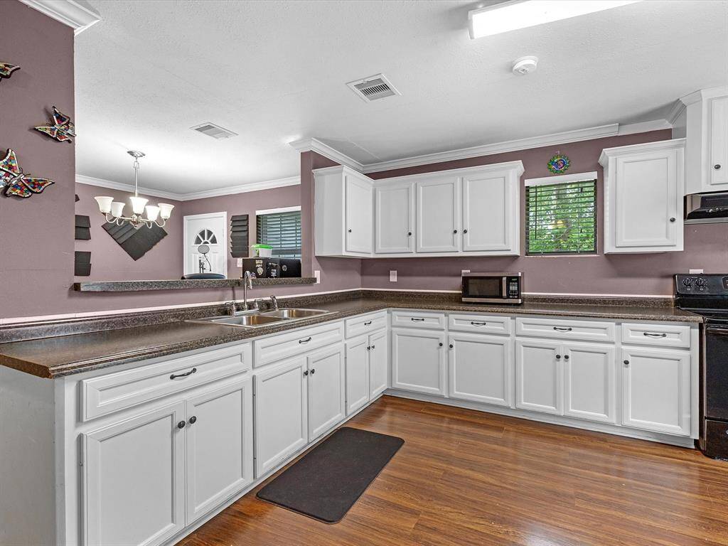 This spacious kitchen features white cabinetry, dark countertops, and wood flooring. It boasts a double sink, ample storage, and a cozy dining area with a chandelier. Natural light enters through a window with blinds, creating a bright and inviting space.