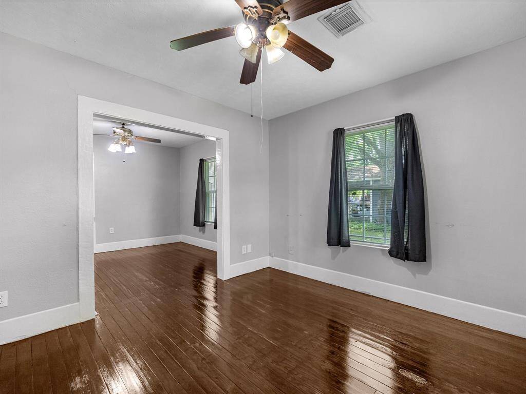 Bright, spacious room with polished wood floors and neutral gray walls. Features ceiling fans, large windows with dark curtains, and an open doorway to an adjacent room.
