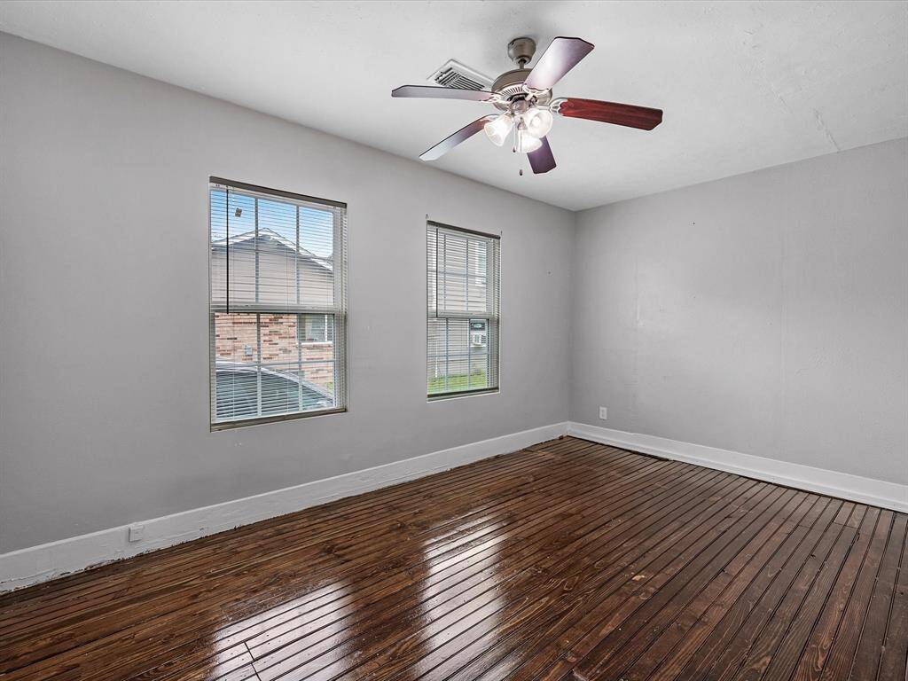 This room features polished wooden floors, two large windows for natural light, neutral gray walls, and a ceiling fan with lights, providing a bright and comfortable space.