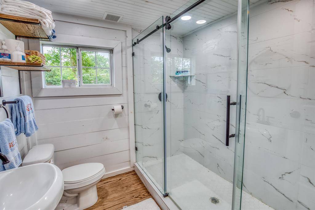 A cozy retreat awaits inside this charming guest suite, featuring warm wood floors and a private bath with a seamless glass-enclosed shower and floor-to-ceiling tile—your own peaceful corner of the property.