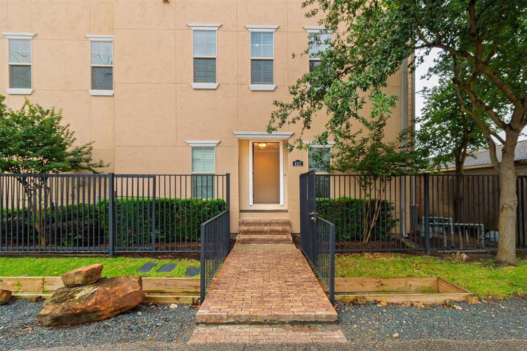 Brick pathway leads you through the front gate. Front patio gate and front door are both open in this picture. Neatly trimmed shrubs and trees add greenery and a sense of privacy. Large and small decorative rocks enhance the landscaping.