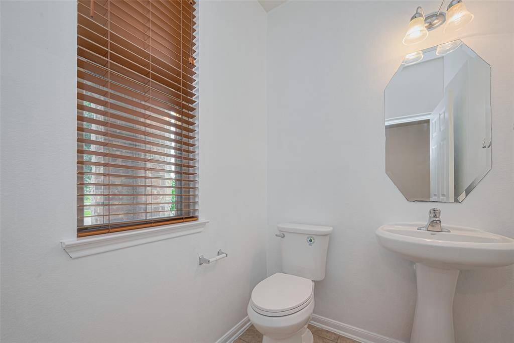 This photo showcases a bright, compact bathroom with a pedestal sink, toilet, and a window with wooden blinds. The space features a wall-mounted mirror and overhead lighting, providing a clean and simple aesthetic.