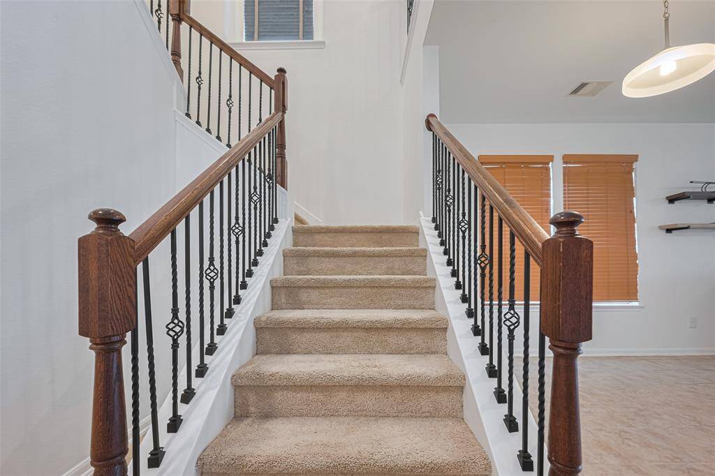A well-lit staircase with beige carpeting and elegant dark wood railings featuring decorative black iron balusters. The space is brightened by natural light from nearby windows, adding warmth to the area.