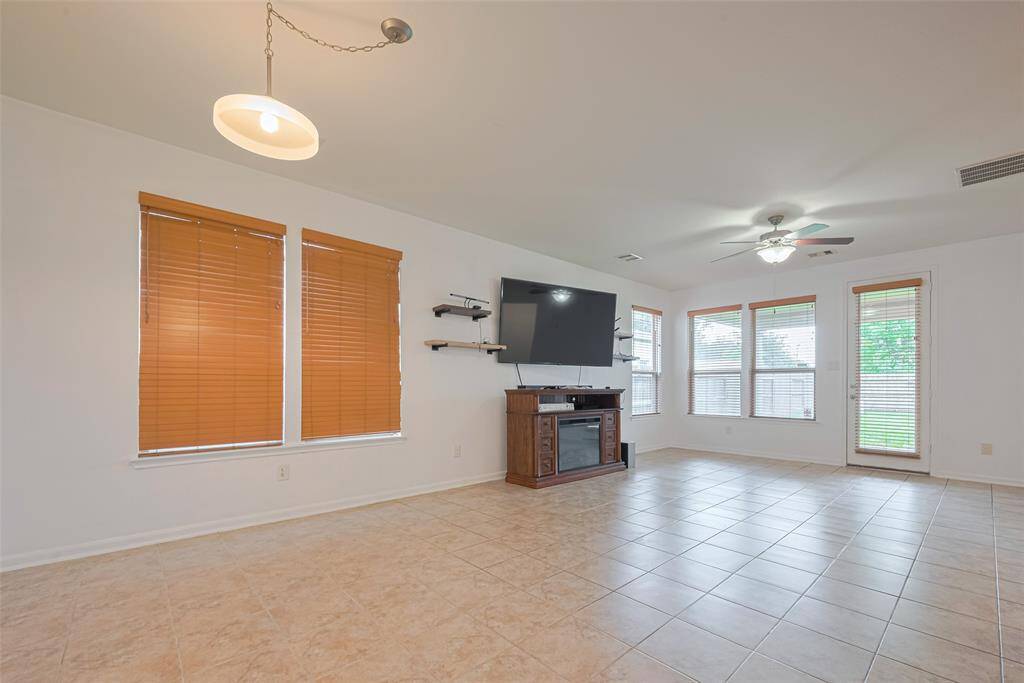 Bright living room with large windows, tiled flooring, and modern light fixtures. Features wall-mounted TV, ceiling fan, and access to the backyard.