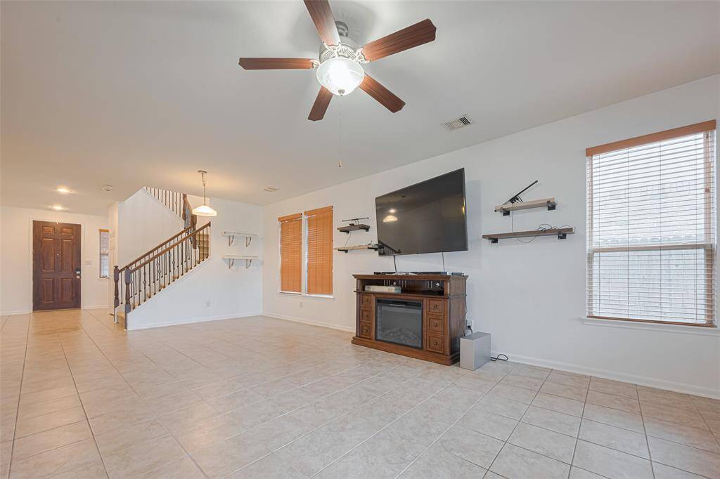Spacious living area with tiled flooring, ceiling fan, and large windows for natural light. Features include a mounted TV, shelves, and a staircase leading to the upper floor. Perfect for a modern and comfortable lifestyle.